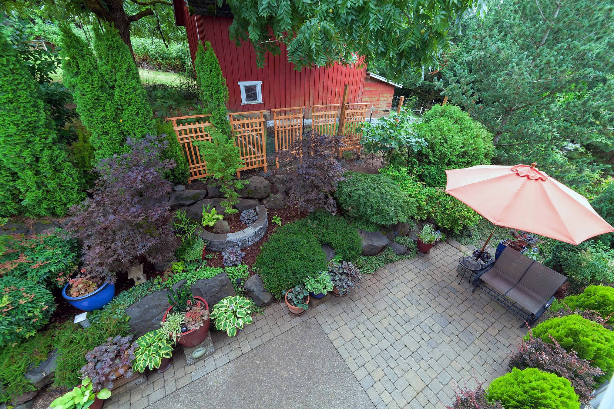 Lush garden featuring diverse plants in pots and a cozy seating area beneath an orange umbrella, set beside a red barn.